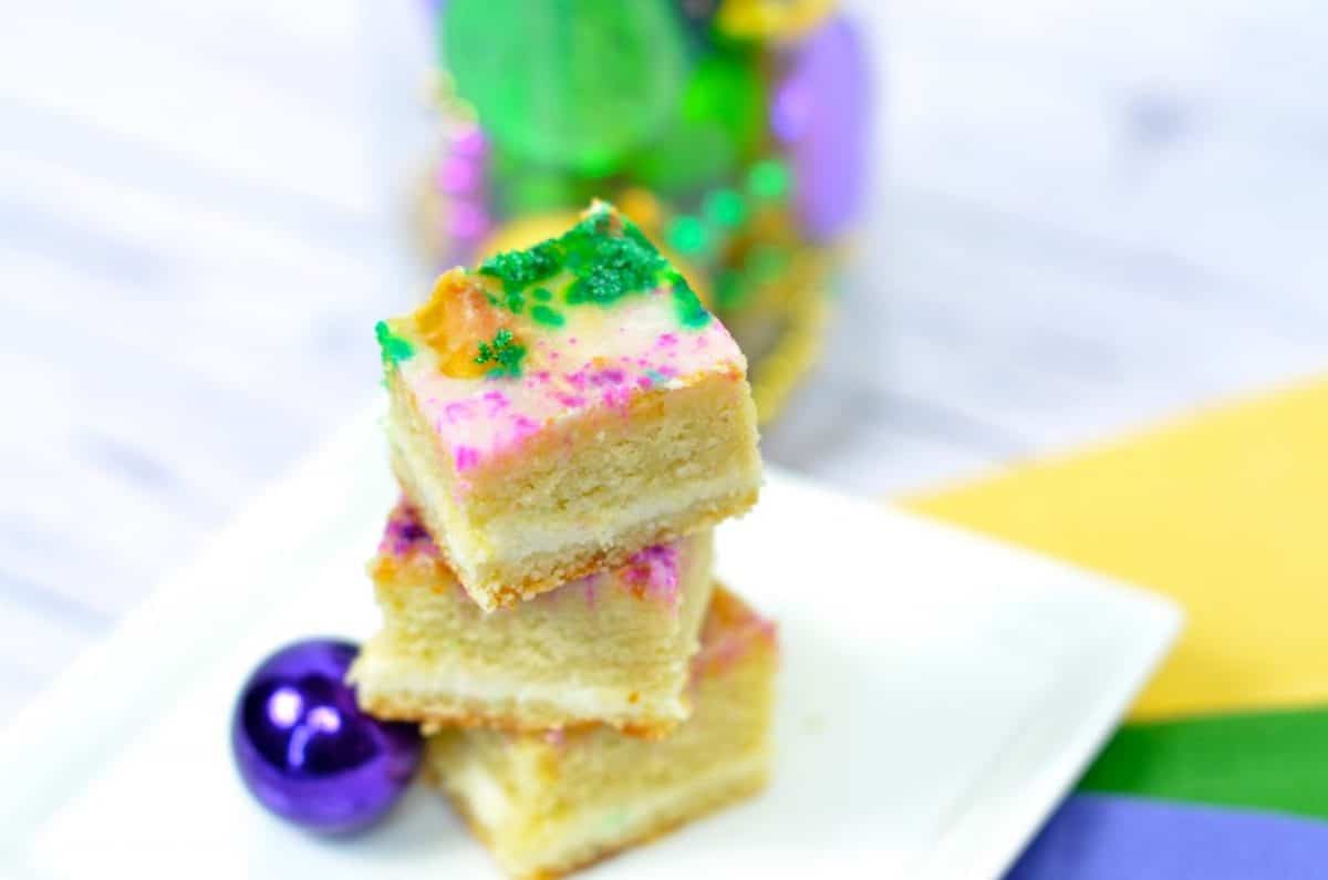 King cake cut into 3 small squares served on a white plate on top of a yellow, green, and purple napkin.