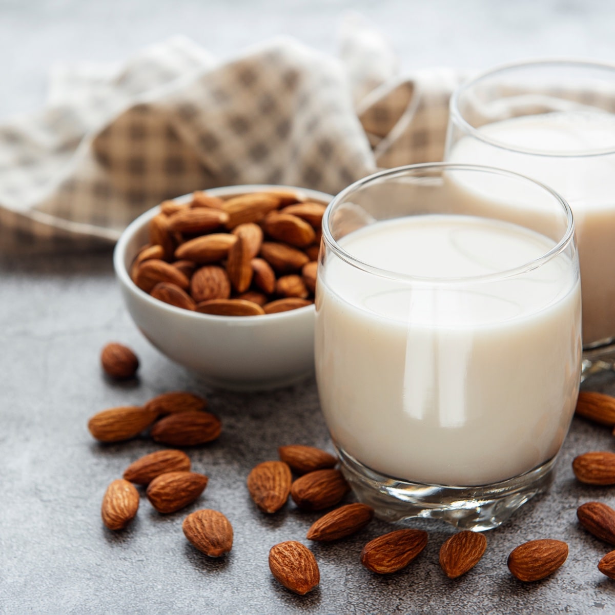 A full glass of almond milk set on a table with a smaller bowl of almond next to the almond milk. 