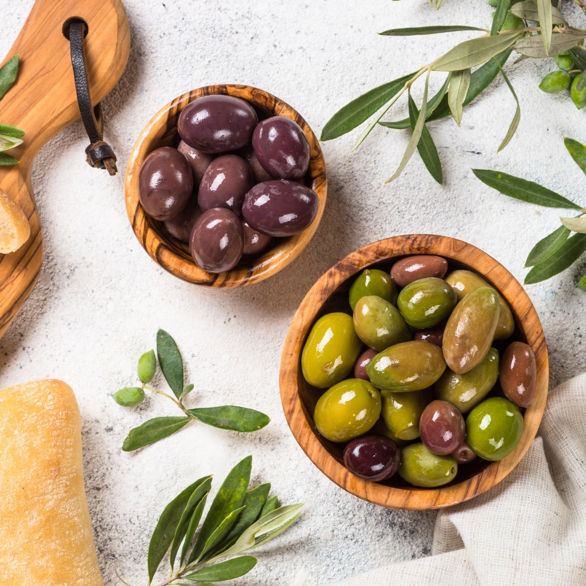 Green olives in a wooden bowl and black olives in a wooden bowl sitting on a white table. 